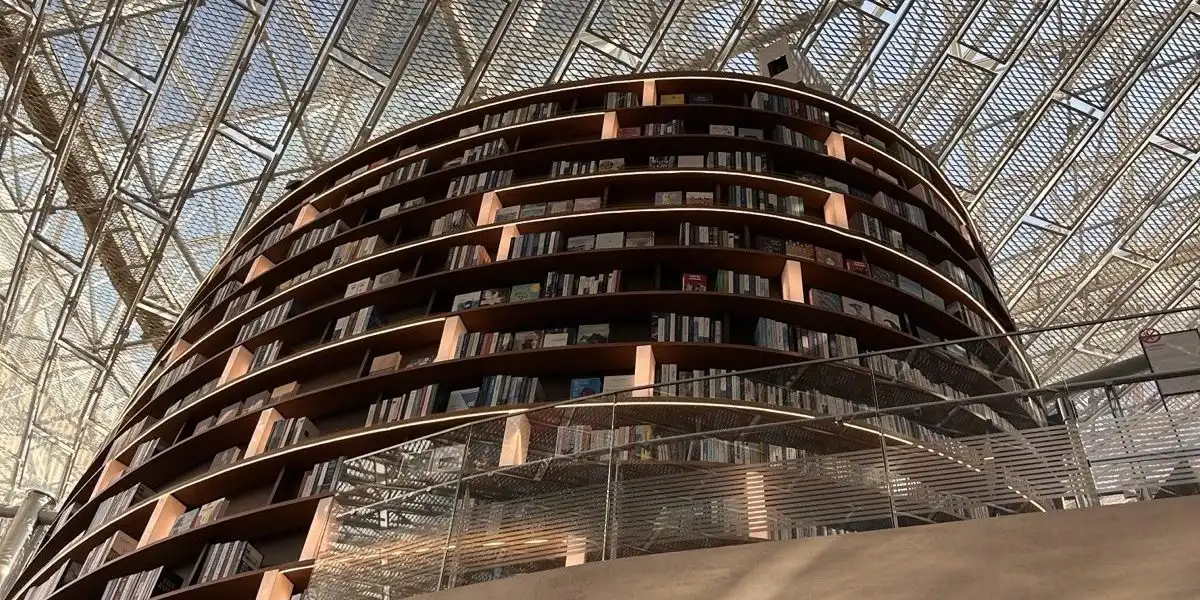 A tall curved bookcase of wooden shelves inside a library, under a lattice roof of glass and steel.