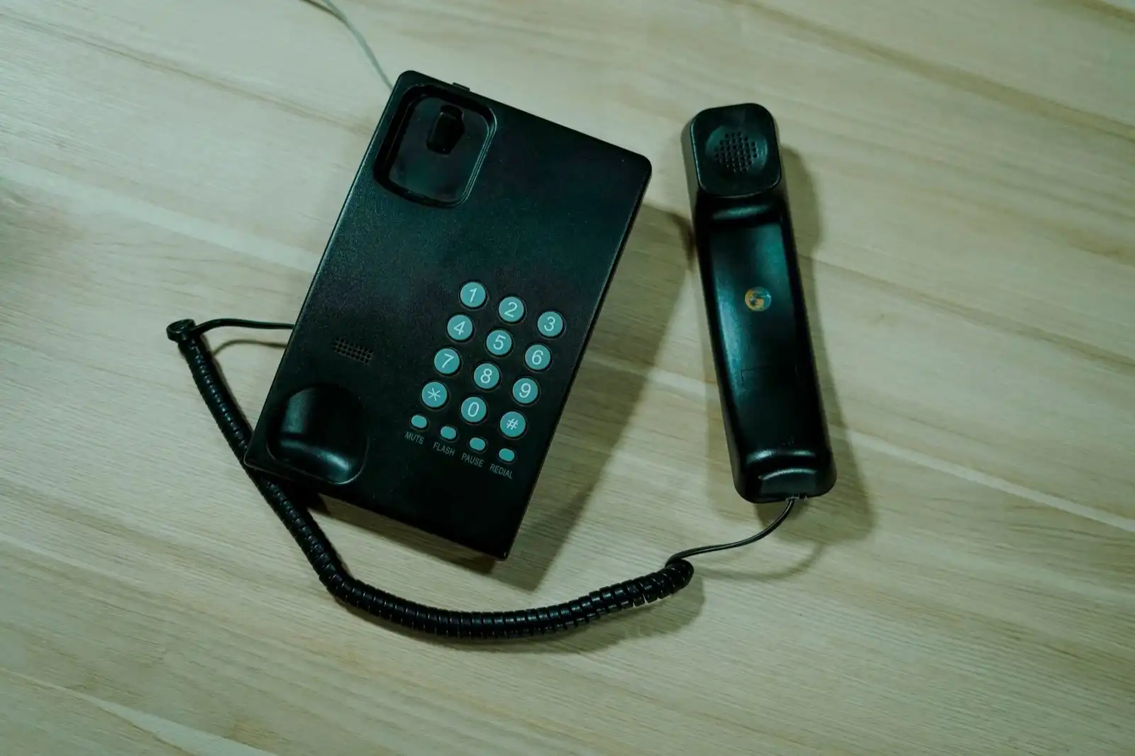 A black push-button desk telephone with its handset lying beside it on a pale wooden table.