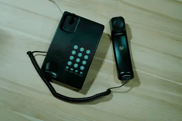 A black push-button desk telephone with its handset lying beside it on a pale wooden table.