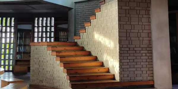 A wooden staircase climbing beside a pale brick wall in a library, with shelves and a lattice window behind.