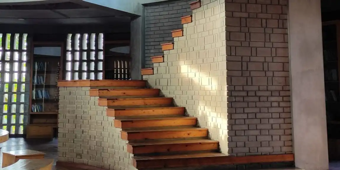 A wooden staircase climbing beside a pale brick wall in a library, with shelves and a lattice window behind.