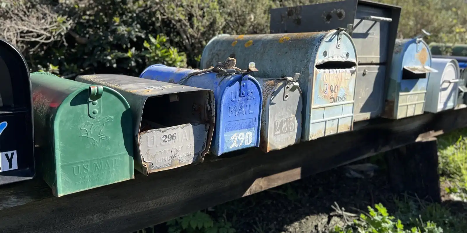 A row of weathered metal mailboxes in green, blue and grey on a long wooden rail, with numbers painted on their doors.