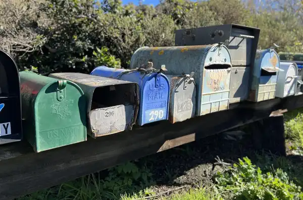 A row of weathered metal mailboxes in green, blue and grey on a long wooden rail, with numbers painted on their doors.