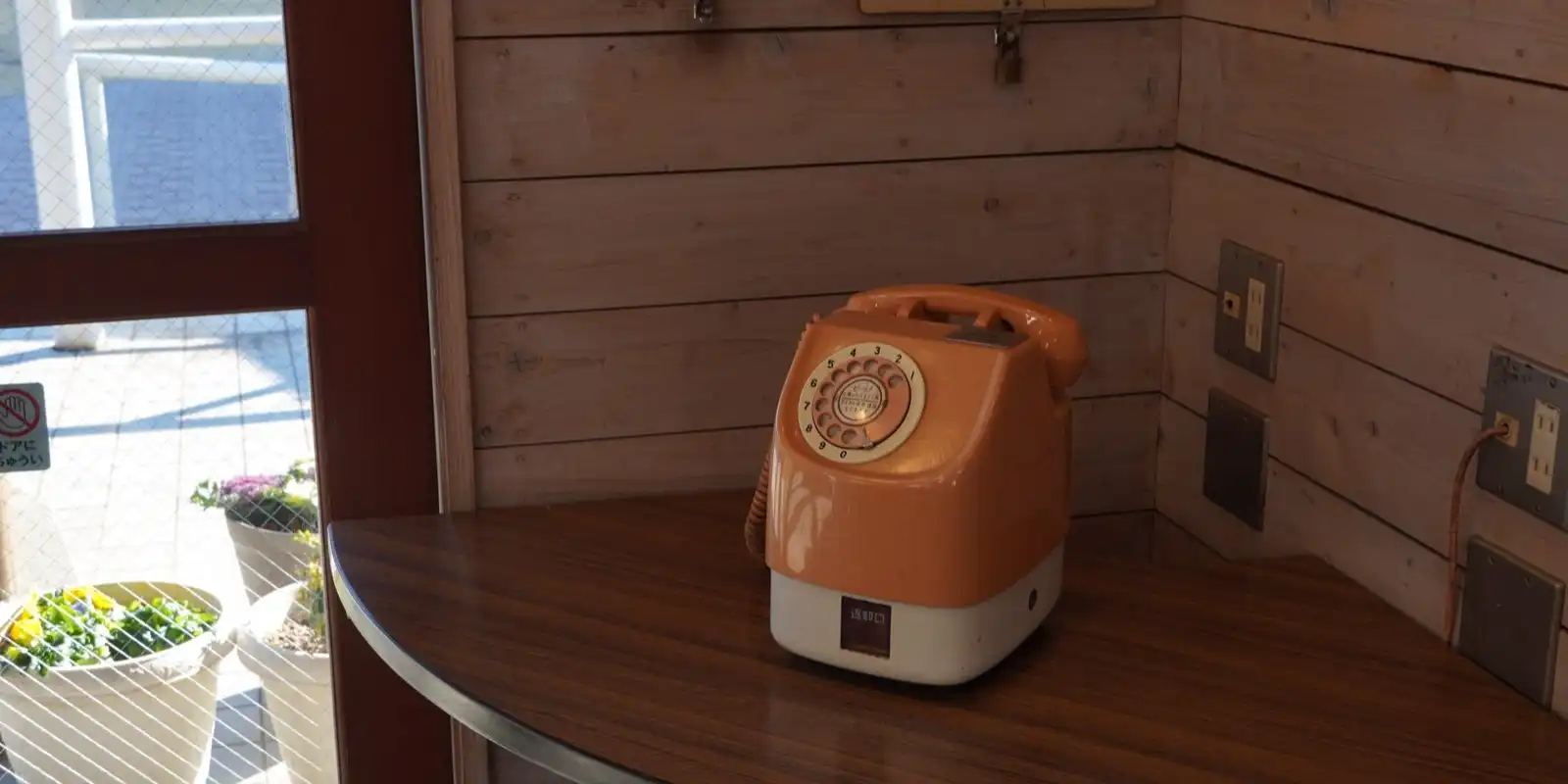 An orange rotary telephone on a curved wooden shelf in the corner of a wood-panelled booth.