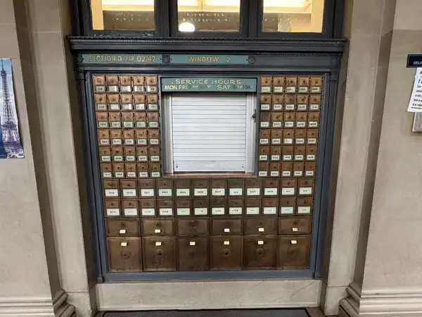 A wall of small numbered metal post office boxes around a closed service window.