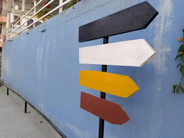 A signpost with four wooden arrows, black, white, yellow and red, in front of a pale blue wall.