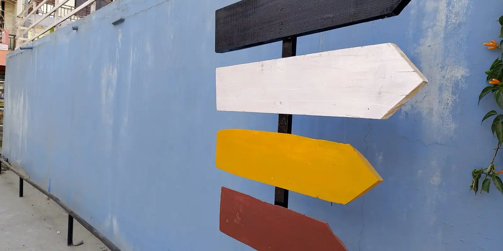 A signpost with four wooden arrows, black, white, yellow and red, in front of a pale blue wall.