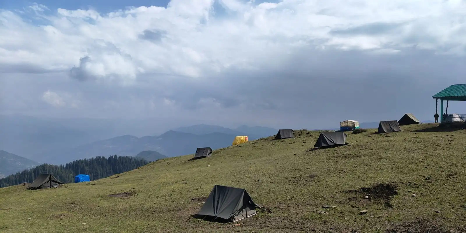 Small dark tents pitched across a grassy ridge, with forested hills and low cloud behind.