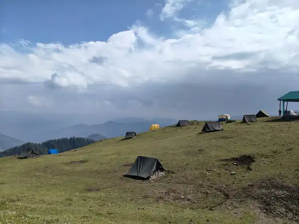 Small dark tents pitched across a grassy ridge, with forested hills and low cloud behind.